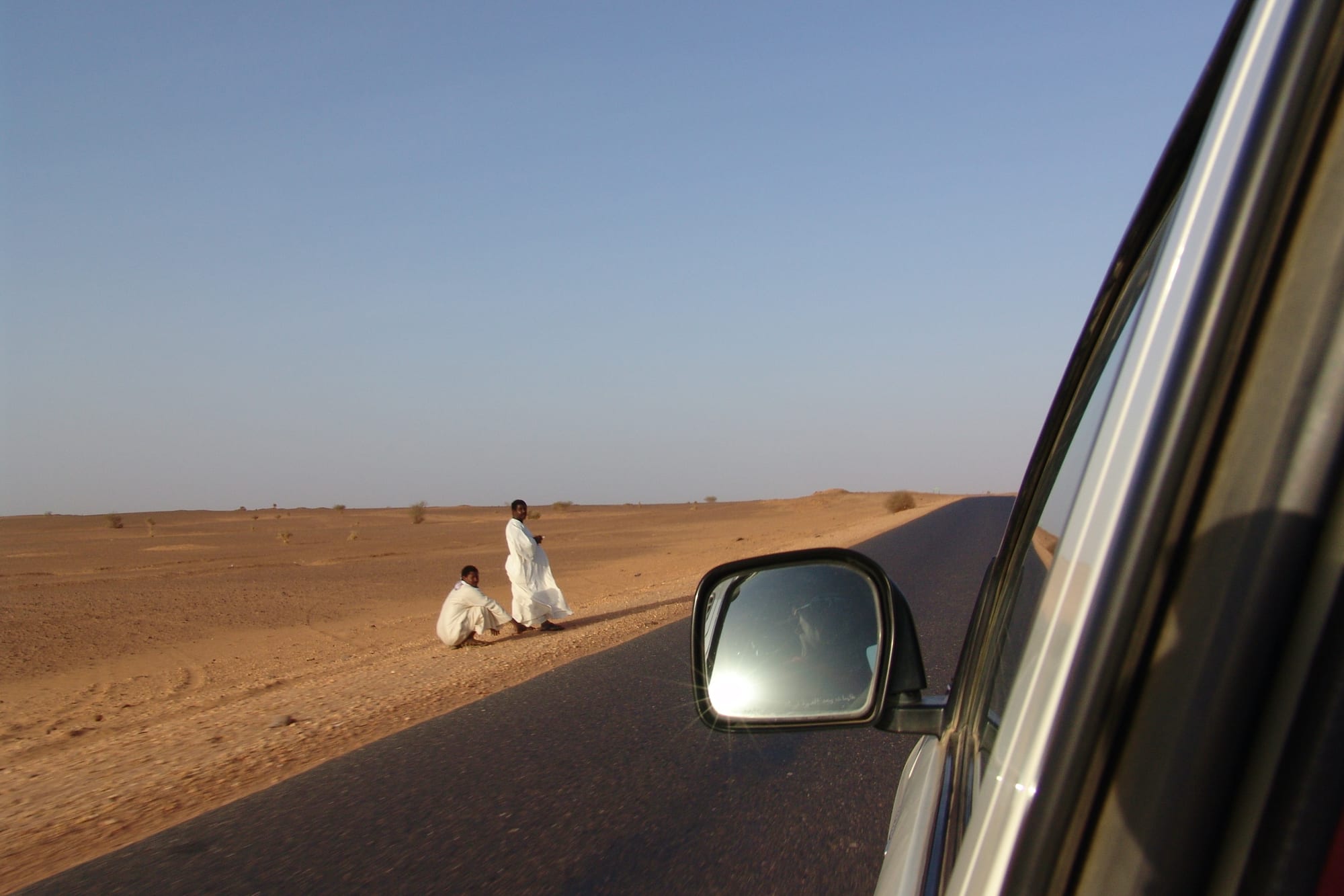 Due uomini seduti lungo la pista nel deserto, visti dallo specchietto della jeep. Sudan