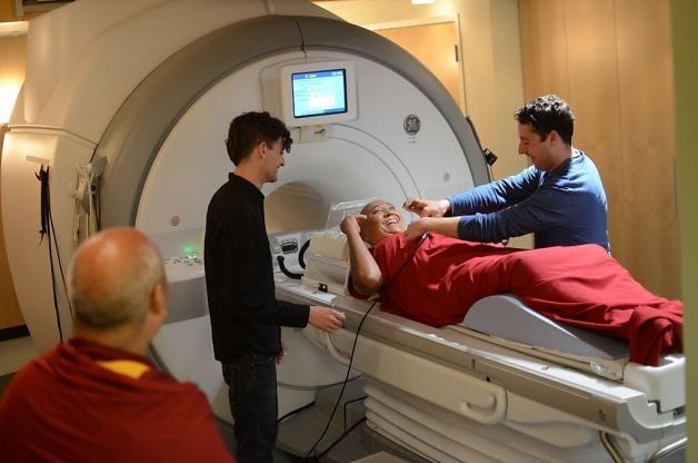 Un monaco disteso e sorridente sul lettino di uno scanner cerebrale, circondato da tre ricercatori durante una pausa dell'esperimento.